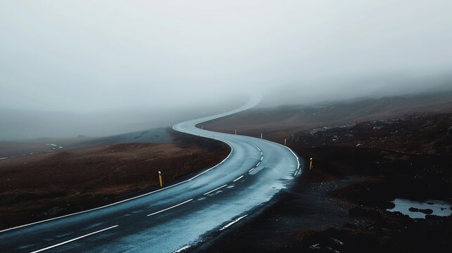 Winding asphalt road disappearing into dense fog and overcast sky over a barren landscape