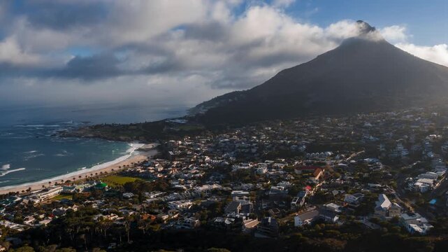 Aerial timelapse shows Lion's Head above Camps Bay in Cape Town. Low clouds wrap the peak as sun and shadows move over rooftops and waves roll onto the beach.
