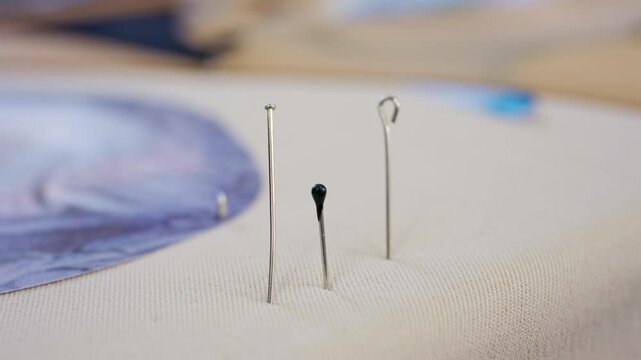 Macro shot of two thin metal sewing pins standing upright in beige woven fabric, with a soft blue paper or fabric pattern blurred in the background