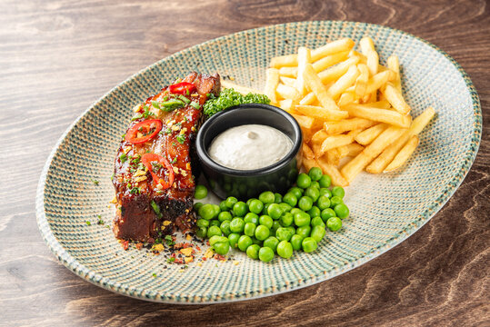 Glazed pork ribs served with french fries, green peas, and dipping sauce on a patterned plate.