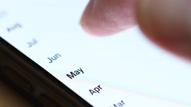 Macro 4K view of a finger scrolling through a vertical list of abbreviated month names on a smartphone screen, with data details blurred on the side.