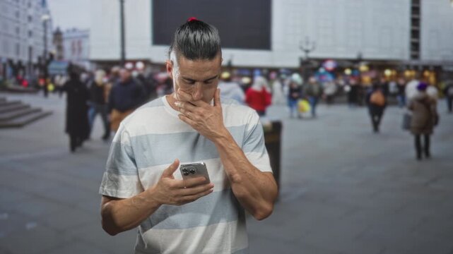 Man holding smartphone with visible hands scrolling and looking down at screen in a crowded street plaza; contemplation.