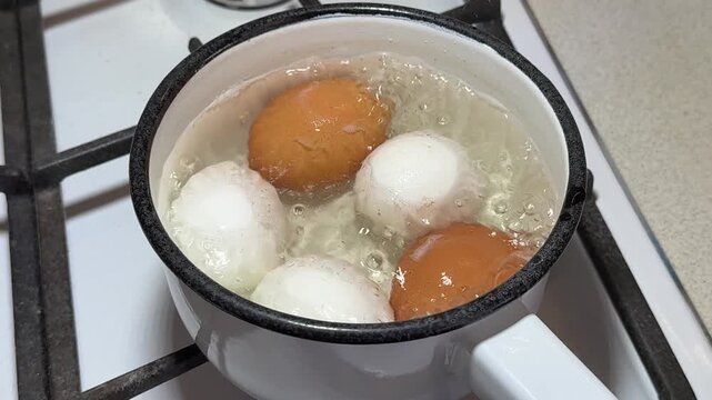 Close-up boiling eggs in enameled ladle on gas stove, brown and white chicken eggs simmering in bubbling water, steam rising, white
