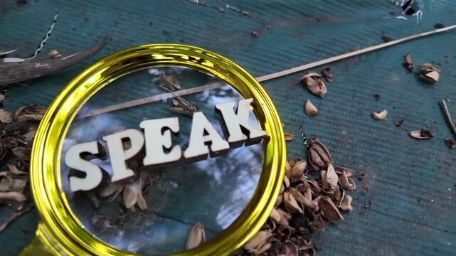 Magnifying glass over wooden letters spelling SPEAK on a textured surface