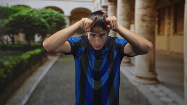 Man soccer player wearing blue striped jersey points both index fingers to his head in building courtyard while squinting and leaning forward; playful taunt.