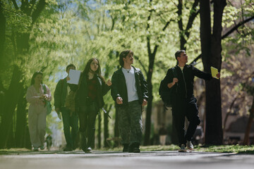 Fototapeta premium A group of school friends walking on a tree-lined campus path carrying backpacks and notebooks. Young students stroll together outdoors on a sunny spring day.