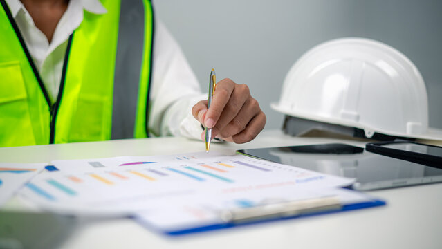 Engineer in safety vest holding pen and analyzing financial graph reports on office desk with white hardhat. Concept of construction management, budget planning, and industrial data analysis.