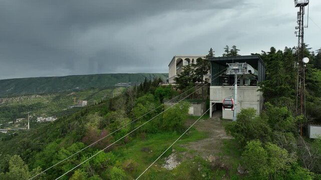 Aerial view of the modern cable car system in Tbilisi, Georgia, showing the upper station building, ropeway cables, and lush green hills under a dark cloudy sky
