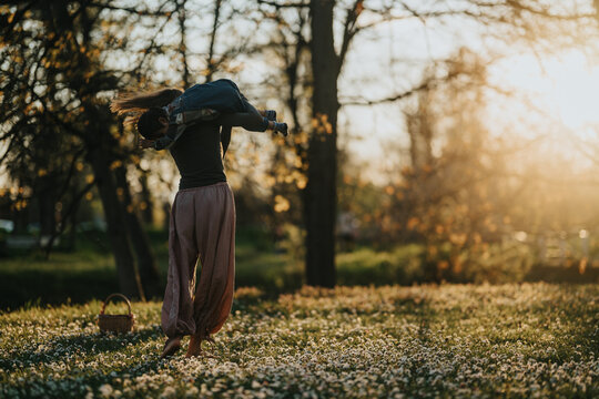 Mother carries her child playfully over her shoulders while walking through a field of small white flowers. Warm golden-hour light filters through trees as they enjoy a quiet outdoor moment together.