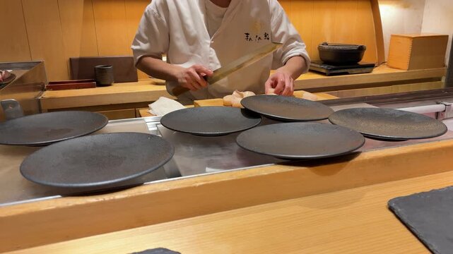 Professional sushi chef prepares fresh raw scallops at an authentic omakase restaurant counter in Tokyo. The master carefully cuts sea mollusk meat using a traditional long Japanese yanagiba knife.