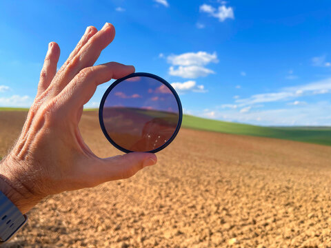 Close up of a photographer's hand holding a round polarizing filter showing the color contrast effect over a sunny plowed field and blue sky background in the countryside
