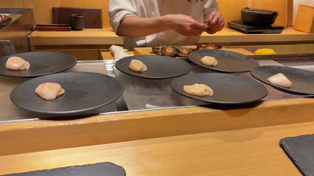 Professional Japanese chef prepping raw seafood on cutting board in Tokyo restaurant. Fresh raw scallop meat served on black plates for traditional omakase course dinner. 
