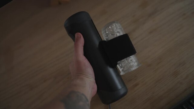 Male hand with a tattoo holding an automatic stroker sex toy over a wooden desk