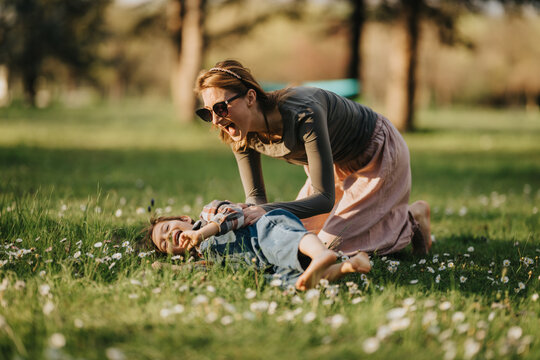 Mother tickling her young daughter on the grass, both laughing joyfully. A playful outdoor moment shows family bonding and carefree fun in a park.