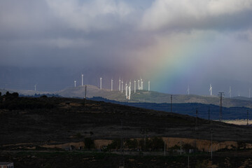 Fototapeta premium Distant view of wind turbines on a hill under a dramatic sky with a bright rainbow, concept of green renewable energy and sustainable environment