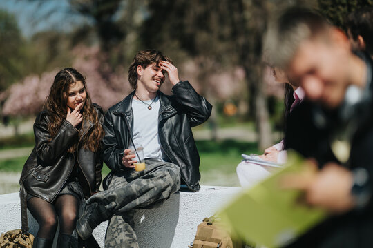 A group of school friends sit together outdoors, laughing and talking on a low campus wall. Casual teenagers in leather jackets enjoy conversation and juice in a candid springtime moment.