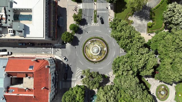 Aerial view of a fountain roundabout with running water feature surrounded by cars trees and terracotta rooftop buildings with a tiered park amphitheater nearby in a Portuguese city