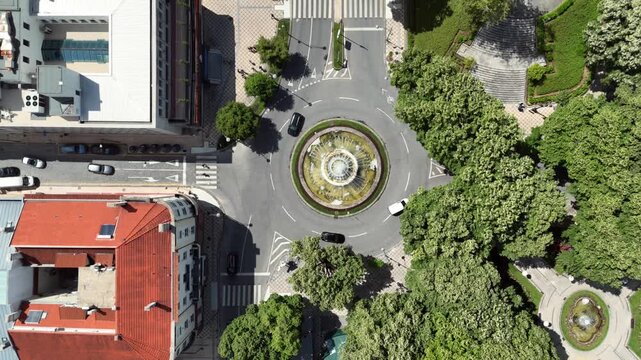 Top down aerial view of a fountain roundabout with active water jets at the center surrounded by vehicles and terracotta rooftop buildings next to a lush green park in Portugal