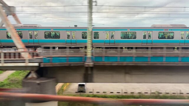 A passenger train passes over a steel truss bridge in Japan. The commuter train moves steadily on a multi-track railway line. High angle view shows suburban infrastructure with green riverbanks.