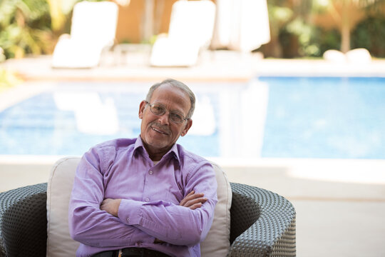 Relaxed senior man sitting poolside on a wicker chair smiling with arms crossed and casual shirt
