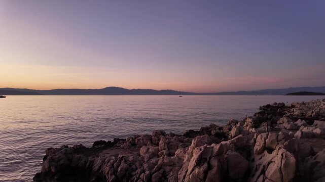 Rocky coastline of the Adriatic Sea near Malinska - Krk Island, Croatia in magical sunset lights. Panning view from the Paradise Coastal Trail or seaside promenade with a fishing boat and wavy water.