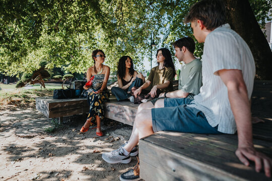 A group of five young friends sit and talk on a wooden bench in a shaded park. Casual conversation and relaxed summer vibes as they gather beneath large leafy trees.