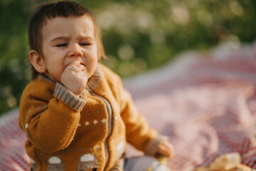 Fototapeta premium A toddler eats a snack while sitting on a picnic blanket in a grassy park. The child in a warm knitted sweater enjoys food outdoors during a casual picnic.