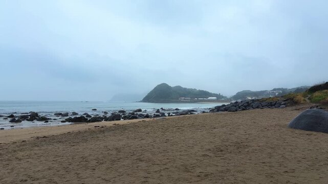 A gloomy view opens up over the sandy and rocky beach in Itoshima, Fukuoka. Dark rocks stretch out into the ocean under a thick blanket of grey clouds. 