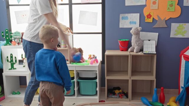 Boy holds blue ball and reaches with hand while woman stands nearby in a playroom building; playful learning.