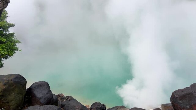 Umi Jigoku hot spring in Japan with thick steam rising from turquoise water in Beppu. Boiling geothermal pond surrounded by rocks and lush green vegetation in Oita Prefecture.