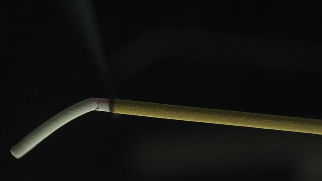 Fragrant incense stick burning and smoking with ash falling against a black background for aromatherapy