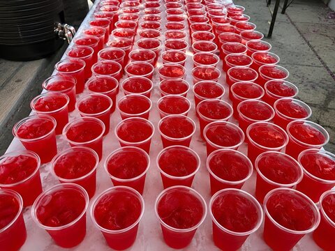 Top view Muharram sabeel stall with red rose sharbat in transparent plastic cups on long white serving table prepared for charitable distribution during Islamic community gathering