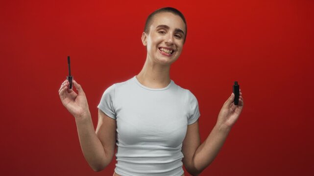 Woman holding mascara wand and tube with raised hands in studio, smiling with short hair and bare hands visible; confidence beauty selfcare.