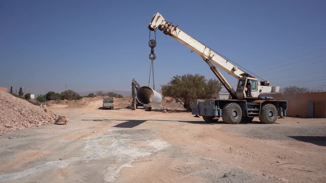 Wide shot of a mobile crane lifting a large pipe at an active construction site, with an excavator working in the background. Essential B-roll for civil engineering and infrastructure projects.