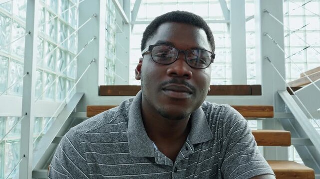 Smiling man adjusts glasses while seated inside modern building with glass walls and wooden stairs
