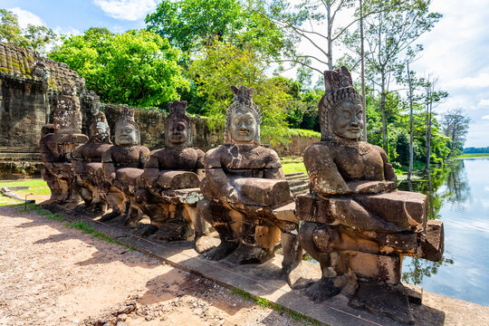 A line of ancient, weathered stone statues of gods or demons stands along a causeway over a moat, leading toward the majestic carved stone entrance gate of angkor thom in Cambodia