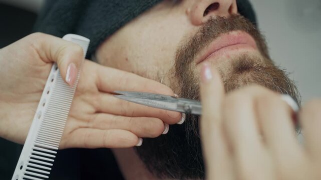Professional barber trimming beard of male client with scissors and comb during grooming procedure in modern barbershop salon