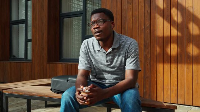 Man sits on bench holding cup while looking outside on a sunny day in a modern urban area with wooden walls