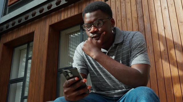 Young man looks at his phone while sitting outside on a wooden bench and enjoying the warm afternoon light