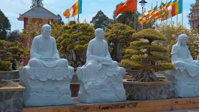 White stone statues of Buddhist monks stand in a row in the garden full of multilayered bonsai trees and Vietnamese National flag together with Buddhist flags flutter on the wind on the background. Vi
