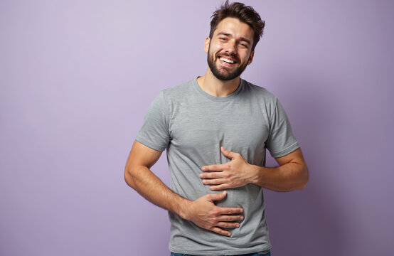 Young man smiles holding belly. Feeling full after meal, possible indigestion or stomach discomfort. Man feels bloated, has belly ache, needs digestive care. Casual t-shirt in studio.