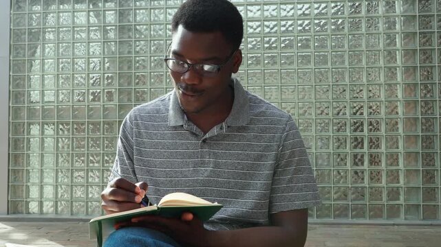 Man writes in notebook while sitting on floor in building with glass wall during afternoon
