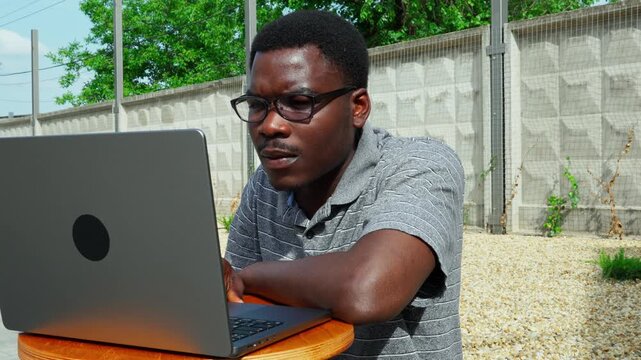Man working on laptop outside in a sunny area with green trees and a wall nearby in the afternoon