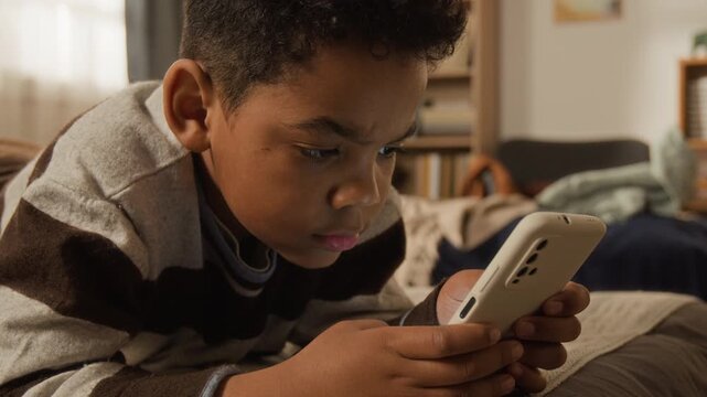 Medium close-up of distracted young 6-year-old black boy lying on bed at home, playing video game on smartphone or browsing social media, representing gadget addiction, screen time and mental health