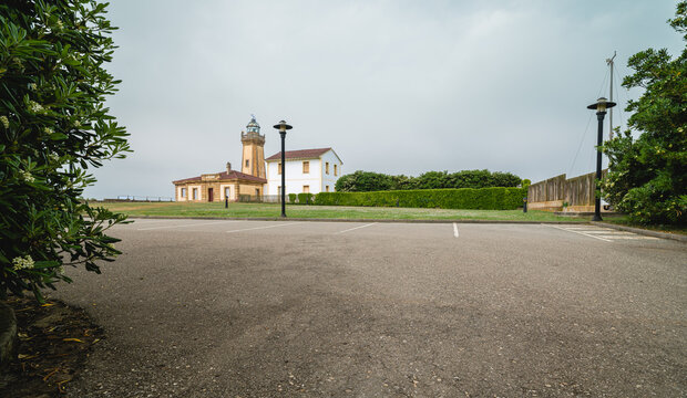 A historic lighthouse and adjacent building, stands prominently on a green lawn overlooking the ocean, with an empty asphalt parking lot in the foreground on an overcast day
