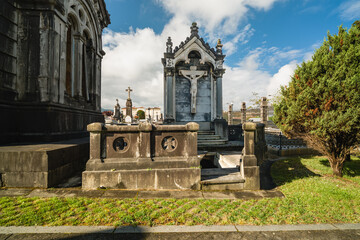 Ornate crucifixion statue on a stone mausoleum among various tombs and gravestones in a historical European cemetery under a blue sky, representing themes of death, religion, and remembrance © Jose Rey