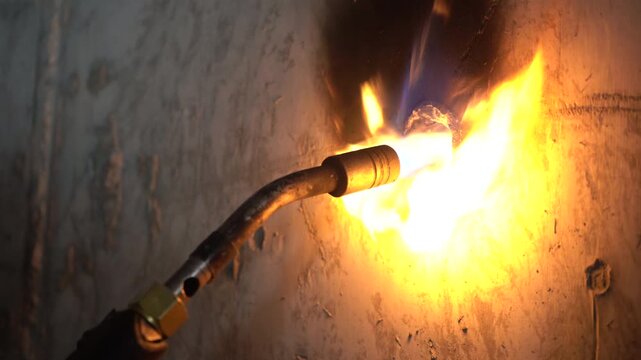 Close-up of a worker heating and scraping a concrete wall penetration with a blowtorch and rebar. Includes authentic roaring and scraping audio. Essential B-roll for civil engineering projects.