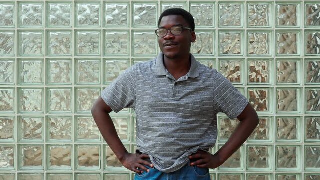 Man stands confidently in front of glass brick wall while wearing casual clothes during a sunny day in the city