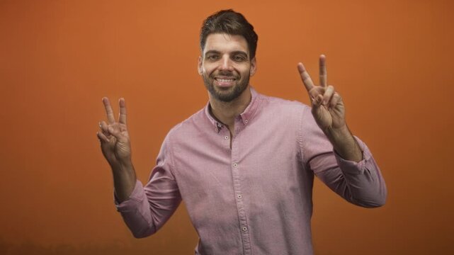 Man with beard wearing a pink shirt shows peace sign with both hands and visible fingers against orange studio backdrop; optimism calm.
