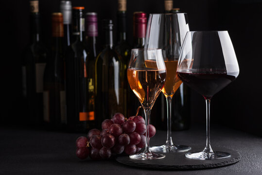 Three glasses of red, white and sparkling rose wine with bunch of grapes on dark slate background